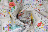 Swirled swatch feed the bees fabric (light grey/white woven look fabric with vertical white rectangle labels/patches with pink, yellow, orange, purple, blue, green floral and tossed bees. Some labels have floral only, some have floral in light blue watering cans, some with yellow beehives)