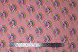 Flat swatch I believe pink fabric (baby pink fabric with repeated rainbow arches with white clouds on either end and "I believe in" text in purple "unicorns" text in rainbow colours)