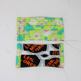 Front and back view of mask with white elastic ear loops (bright neon green mask with drawing style daisies in white and green/blue with long stems and pink centers of white floral)