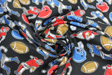 Swirled swatch of football printed fabric on black (black fabric with tossed red and blue football helmets, brown footballs, small cartoon blue and red football players in various tackle/run positions)