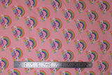 Flat swatch I believe pink fabric (baby pink fabric with repeated rainbow arches with white clouds on either end and "I believe in" text in purple "unicorns" text in rainbow colours)