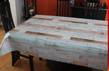 Rustic Wood (weathered boards from off-white to dark tan in offset rows running parallel to the natural edge) opaque vinyl draped over a dining room table with matching chairs around it.
