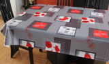 Grey w/poppies (white blocks with red poppies and dark grey borders are scattered among dark grey rectangles and red rectangles with line drawings of poppies in white or red over top, all on a mid grey background) opaque vinyl draped over a dining room table with matching chairs around it.